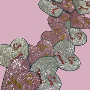 May include: A collection of pink and white heart-shaped patches with sparkly sequins and gold lettering. The patches feature the word "Sister" in different fonts and styles.