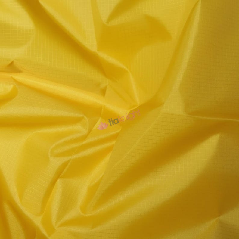 May include: Close-up of a bright yellow fabric with a subtle grid pattern. The material is smooth and appears to be lightweight, with folds and creases adding texture. The overall impression is of a vibrant, sunny textile.