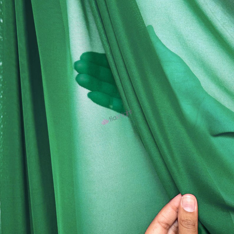 May include: A sheer, emerald green fabric with a fine mesh texture. The material is draped in soft folds, creating varying levels of opacity. The lightweight textile is translucent, allowing light to pass through and revealing a subtle texture.
