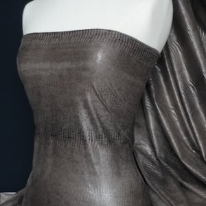 Op de afbeelding: Een close-up van een bruine kunstlederen stof met een subtiel reliëfpatroon van krokodilhuid. De stof heeft een lichte glans en is gedrapeerd over een wit mannequin.