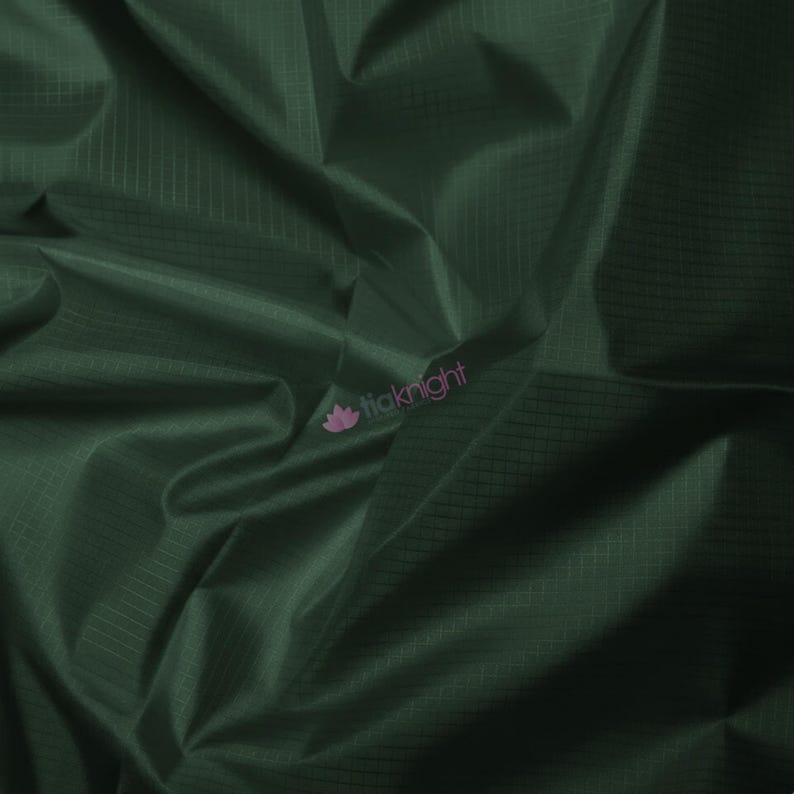 May include: Close-up of a dark green fabric with a subtle grid pattern. The material is folded, creating shadows and highlights that emphasize its texture. The fabric has a slight sheen, suggesting a smooth surface. The logo "tiaknight" is visible in the lower right corner.