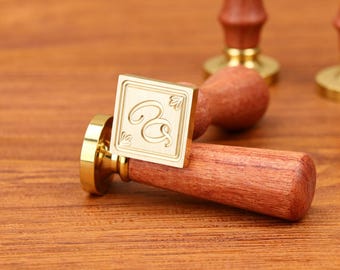 Può includere: Un timbro sigillo di cera color oro con manico in legno. Il timbro quadrato presenta la lettera "R" in una scrittura elegante. Il manico è di un ricco colore marrone. Il timbro è appoggiato su una superficie di legno.