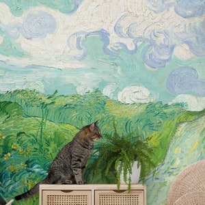 Puede incluir: Un gato atigrado se sienta en un gabinete de madera con dos cajones de mimbre. El gabinete está frente a una pared con un mural de paisaje verde y azul. Una planta de helecho en maceta está en el gabinete al lado del gato.