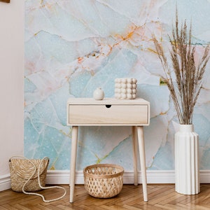 May include: A light wood nightstand with a drawer and tapered legs. The nightstand is in front of a wall with a light blue and pink marble wallpaper. A small white ceramic vase with a white ceramic orb sits on top of the nightstand. A woven basket sits on the floor in front of the nightstand. A white ceramic vase with a dried grass arrangement sits on the floor to the right of the nightstand.
