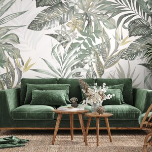May include: A green velvet sofa with wooden legs sits in a living room with a tropical leaf patterned wallpaper. Two wooden coffee tables and a wicker chair are also in the room.