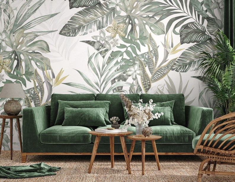 May include: A green velvet sofa with wooden legs sits in a living room with a tropical leaf patterned wallpaper. Two wooden coffee tables and a wicker chair are also in the room.
