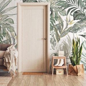May include: A white wall with a green tropical leaf wallpaper pattern. A brown armchair with a white throw blanket is in the foreground. A wooden door is to the left of the image. A small wooden stool with a white box and a pair of white shoes is to the right of the image. A potted plant with a brown burlap bag is in the right corner of the image.