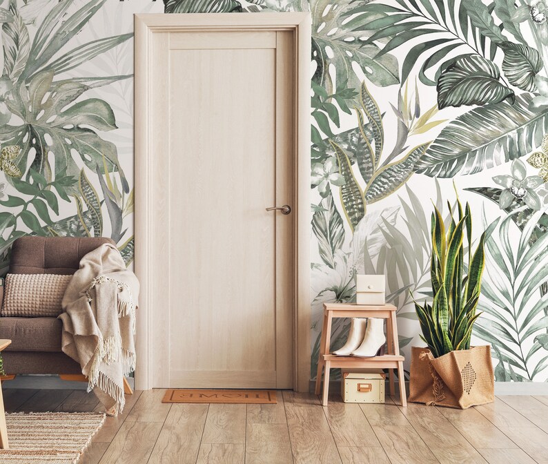 May include: A white wall with a green tropical leaf wallpaper pattern. A brown armchair with a white throw blanket is in the foreground. A wooden door is to the left of the image. A small wooden stool with a white box and a pair of white shoes is to the right of the image. A potted plant with a brown burlap bag is in the right corner of the image.