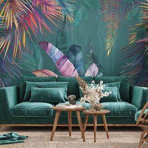 May include: A teal velvet sofa with two pillows sits in front of a wall with a tropical leaf pattern. The leaves are painted in shades of pink, purple, and green. There are two wooden coffee tables in front of the sofa and a wicker chair to the right.