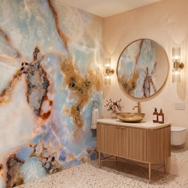 May include: A bathroom vanity featuring a fluted wooden cabinet with a brass base, a gold vessel sink, and a round mirror. The wall is covered in large-scale blue and amber marble slabs, complemented by two vertical glass wall sconces and a towel.