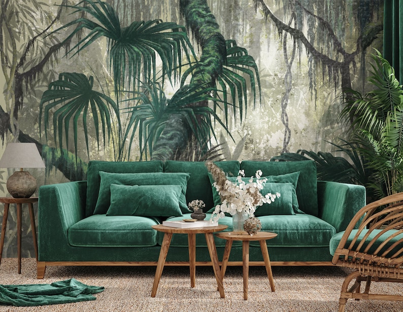 Puede incluir: Un sof&aacute; de terciopelo verde con dos almohadas se encuentra frente a un mural de pared que representa una exuberante escena de jungla tropical. El sof&aacute; est&aacute; acentuado con dos mesas de caf&eacute; de madera y una silla de mimbre.