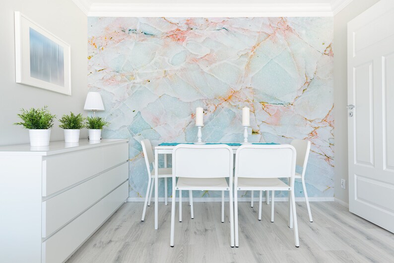 May include: A white dining table with four white chairs and a blue runner. The wall behind the table is covered in a marble-patterned wallpaper with pink, blue, and gold accents.