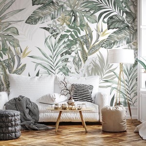 May include: A white sofa with a gray throw blanket and a beige pouf in a living room with a tropical leaf patterned wallpaper.