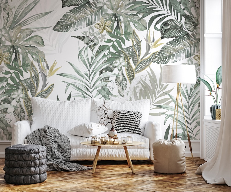 May include: A white sofa with a gray throw blanket and a beige pouf in a living room with a tropical leaf patterned wallpaper.