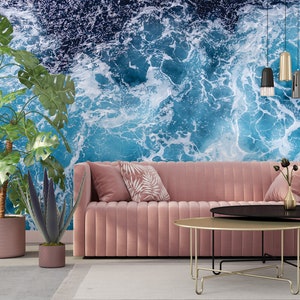 Może przedstawiać: R&oacute;żowa welurowa sofa z dwoma poduszkami i złotym metalowym stolikiem kawowym przed nią. Ściana za sofą to mural z niebieskimi falami oceanu.