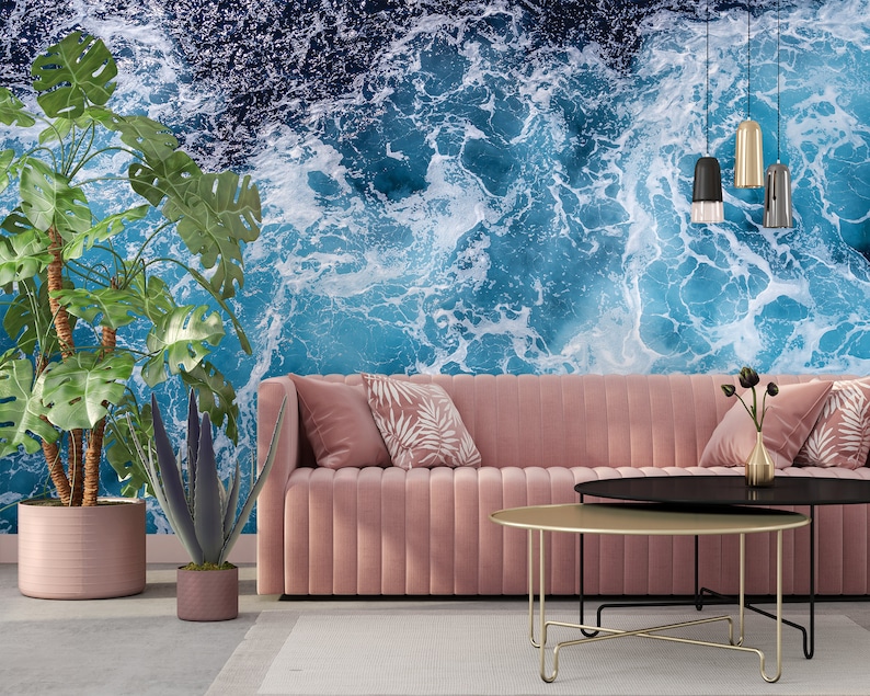 Może przedstawiać: R&oacute;żowa welurowa sofa z dwoma poduszkami i złotym metalowym stolikiem kawowym przed nią. Ściana za sofą to mural z niebieskimi falami oceanu.