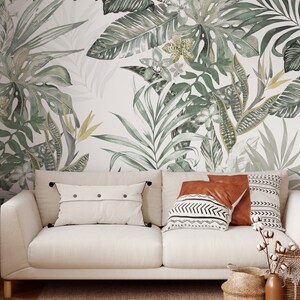 May include: A white sofa with a light brown throw pillow, a brown throw pillow with a black and white geometric pattern, and a white throw pillow with black buttons. The sofa is in front of a wall with a green and white tropical leaf wallpaper.