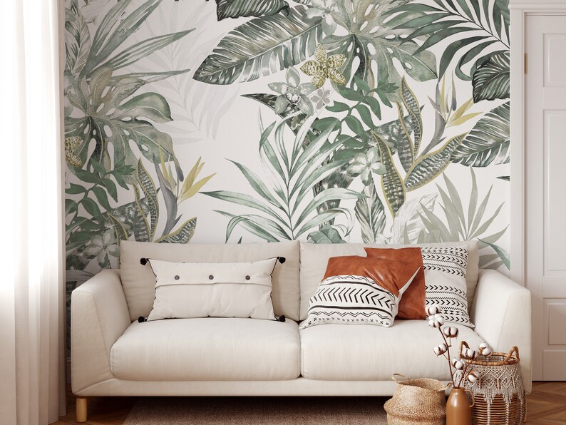 May include: A white sofa with a light brown throw pillow, a brown throw pillow with a black and white geometric pattern, and a white throw pillow with black buttons. The sofa is in front of a wall with a green and white tropical leaf wallpaper.