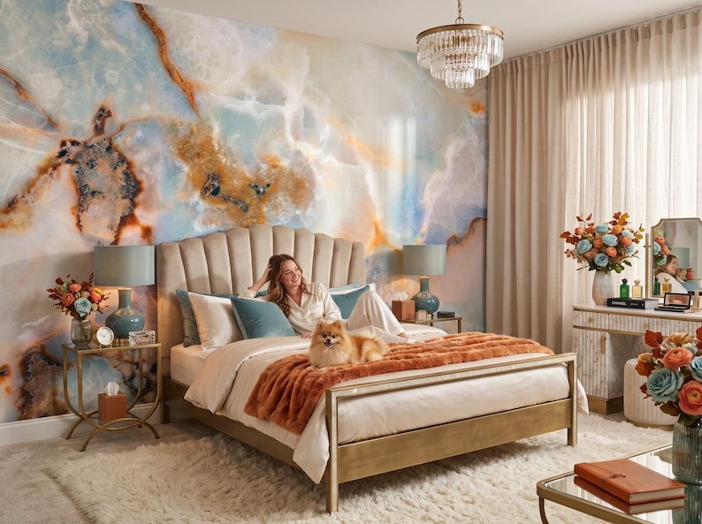 May include: A bedroom featuring a scalloped beige upholstered bed frame with a plush orange throw blanket, set against a large-scale wall mural with blue, white, and amber marble patterns, complemented by a crystal chandelier and two bedside lamps.