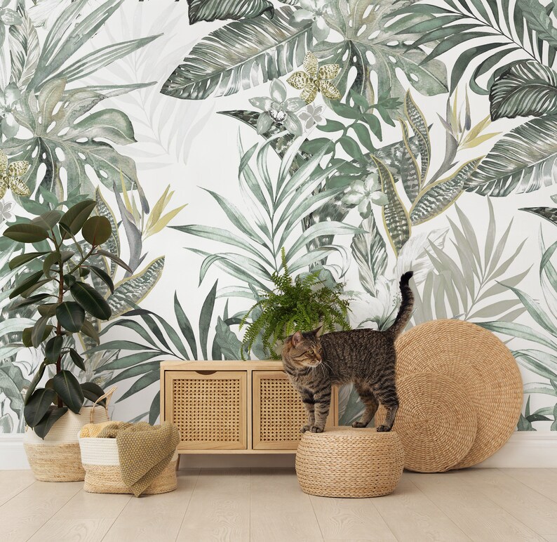 May include: A tabby cat sits on a woven stool in front of a wooden cabinet with woven doors. The cat is looking to the left. The background is a white wall with a green and white tropical leaf pattern.