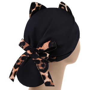May include: Black scrub cap with cat ears and a bow. The cap features a leopard print on the ears and bow, contrasting with the solid black fabric of the cap. The bow is tied at the back.