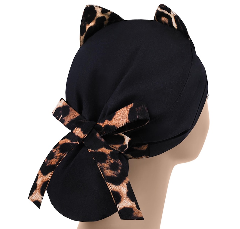 May include: Black scrub cap with cat ears and a bow. The cap features a leopard print on the ears and bow, contrasting with the solid black fabric of the cap. The bow is tied at the back.