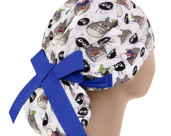 May include: White scrub cap featuring a repeating pattern of cartoon characters in gray, black, and green. The cap has a blue bow on the side. The cap is designed to be worn in a medical or healthcare setting.