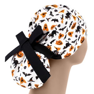 Puede incluir:  gorro de quirófano blanco con un lazo negro y un estampado de Halloween con calabazas naranjas, gatos negros, murciélagos y fantasmas.