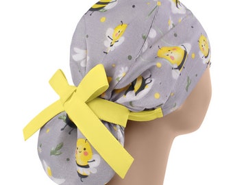 May include: A gray scrub cap featuring a repeating pattern of yellow cartoon bees and white daisies. The cap has a yellow bow and trim. The cap is designed to be worn on the head.