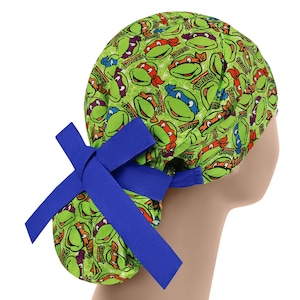 Puede incluir: Un gorro quirúrgico verde con un patrón repetido de caras de las Tortugas Ninja en rojo, azul y morado. El gorro tiene un lazo azul en el lateral. El fondo es blanco.