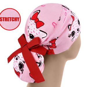 Könnte beinhalten: Eine rosa OP-Haube mit einem wiederkehrenden Muster aus weißen Katzengesichtern mit roten Schleifen. Die Haube verfügt über ein rotes Stoffband am Hinterkopf, um den Pferdeschwanzbeutel zu fixieren. Ein roter Kreis enthält den Text STRETCHY.