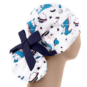 May include: White surgical scrub hat with a blue and red dinosaur and spaceship print. The hat has a navy blue bow tie.
