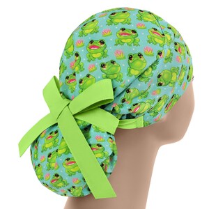 Pode incluir: Uma touca cirúrgica azul clara com um padrão repetido de sapos verdes sorrindo e nenúfares cor-de-rosa. A touca tem um laço verde limão na parte de trás. Esta touca é projetada para profissionais médicos.