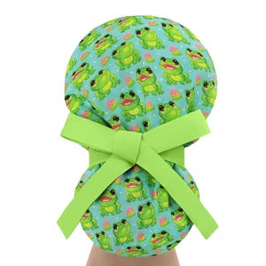 May include: A light green scrub cap with a repeating pattern of smiling green frogs and pink water lilies on a light blue background. The cap has a bright green bow.