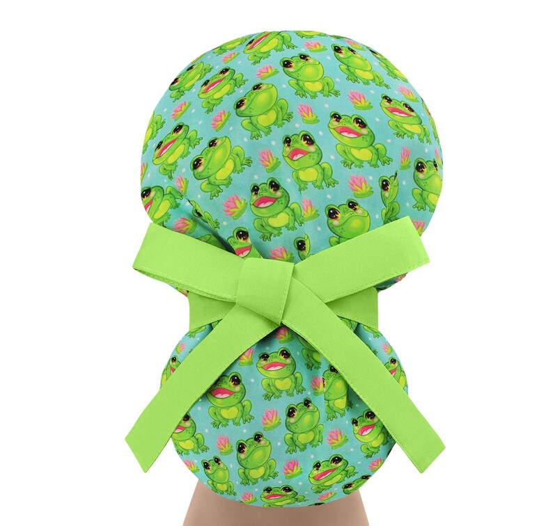 May include: A light green scrub cap with a repeating pattern of smiling green frogs and pink water lilies on a light blue background. The cap has a bright green bow.