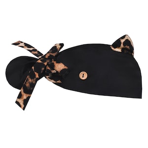 May include: Black scrub cap with a leopard print bow and cat ears. The cap features a wooden button detail and is designed for medical professionals or those in the beauty industry. The leopard print adds a stylish touch to the functional headwear.