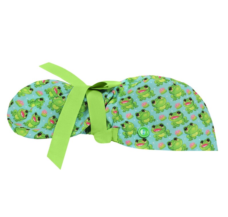 May include: A light blue scrub cap featuring a repeating pattern of smiling green frogs and pink water lilies. The cap has a lime green ribbon for tying and a green button detail.