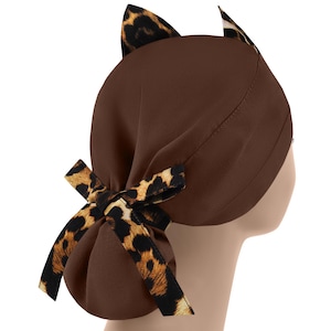 May include: A brown scrub cap with cat ears and a bow. The cap features a leopard print pattern on the ears and bow. The cap is designed to be worn on the head.