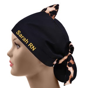 May include: Black scrub cap with a leopard print accent and cat ears. The cap features the text "Sarah RN" in gold lettering. The cap is tied in a bow at the back.