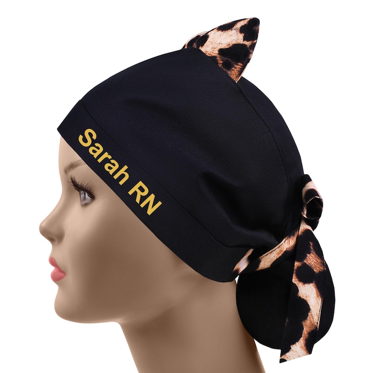 May include: Black scrub cap with a leopard print accent and cat ears. The cap features the text "Sarah RN" in gold lettering. The cap is tied in a bow at the back.