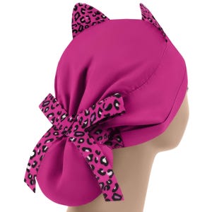 May include: A magenta-colored scrub cap with cat ears and a bow. The ears and bow feature a black and white leopard print pattern. The cap is designed to be worn on the head.