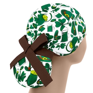 Può includere: Un berretto da lavoro bianco con una stampa verde, gialla e marrone di trifogli, boccali di birra e cappelli da folletto. Il berretto ha un nastro marrone legato sul retro.