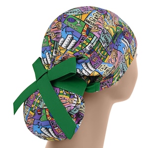 Puede incluir: Un gorro quirúrgico colorido con un lazo verde. El gorro presenta un patrón vibrante con texto e ilustraciones en varios colores. El texto incluye frases como "Keep Calm" y "O₂ To Go".