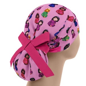 May include: A pink scrub cap featuring a pattern of pregnant woman emojis in various colors. The cap has a large, bright pink bow on the back. The cap is designed to be worn in a medical setting.