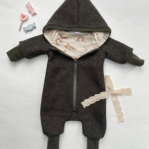 Könnte beinhalten: Ein dunkelgrüner Baby-Overall mit Kapuze und Reißverschluss vorne. Die Kapuze ist mit einem cremefarbenen Stoff mit braunem Fuchs-Druck gefüttert. Der Overall hat einen weißen Spitzenbesatz am Bein.