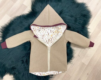 May include: A tan hooded jacket with a full-length zipper. The jacket has a burgundy trim on the hood and cuffs. The interior lining features a floral pattern with white, yellow, and brown flowers on a white background. The jacket is made of a soft, textured fabric.