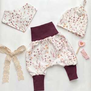 Puede incluir: Un conjunto de ropa de bebé con estampado floral. El conjunto incluye un pantalón blanco y burdeos, un babero blanco con estampado floral y un gorro blanco con estampado floral.