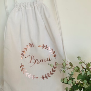 Może przedstawiać: Biała torba ze sznurkiem z różowo-złotym wieńcem kwiatowym i napisem "Braut" w środku.