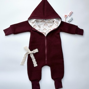 Könnte beinhalten: Ein burgunderfarbener Kapuzenoverall mit einem weißen und braunen Regenbogenmuster, das die Kapuze auskleidet. Der Overall hat einen Reißverschluss vorne und Bündchen an den Beinen.