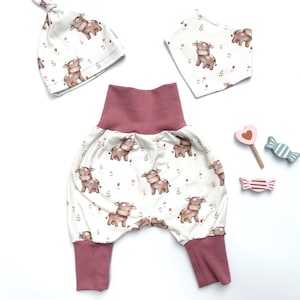 Puede incluir: Un conjunto de ropa para bebé con un gorro blanco, un babero y pantalones con estampado de vaca marrón. El gorro tiene un nudo en la parte superior. Los pantalones tienen una cintura elástica y puños de color rosa.